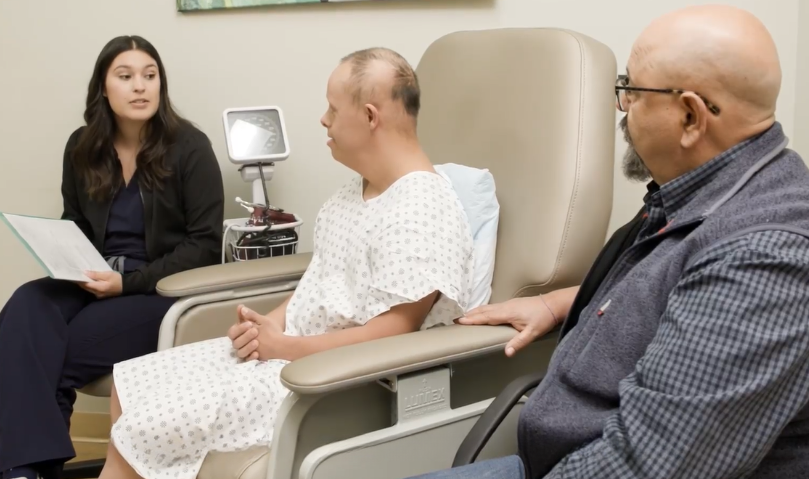 A white man with Down syndrome is seated in an exam chair wearing a hospital gown. Next to him on one side is a young woman with dark hair who is seated and is speaking with papers in her hand and on the other side is a bald man with glasses and a gotee. The men appear to be listening to what the woman is explaining to them.  