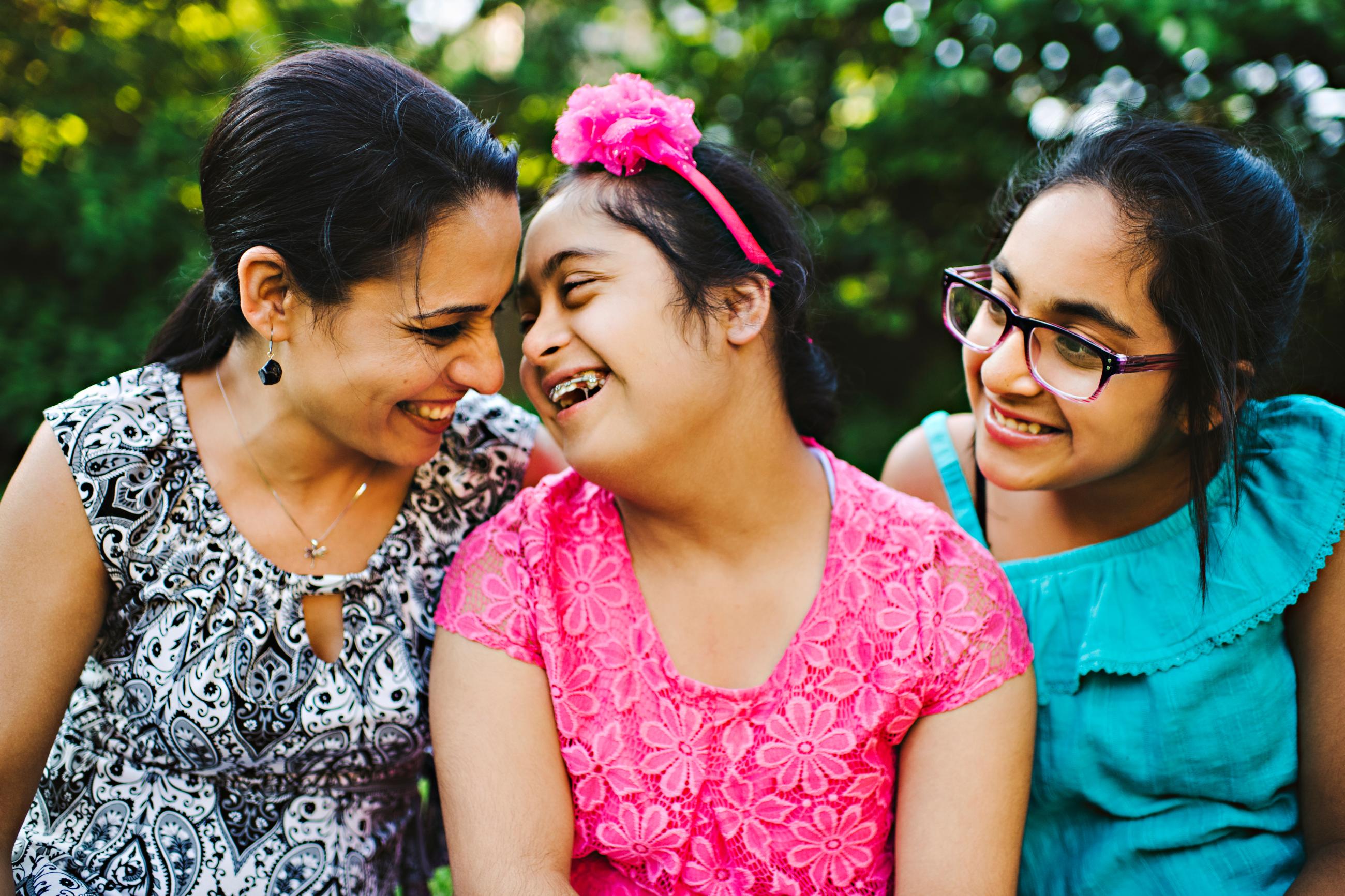 A young woman with Down syndrome smiles with her family. She wears a bright pink shirt and matching floral headband.
