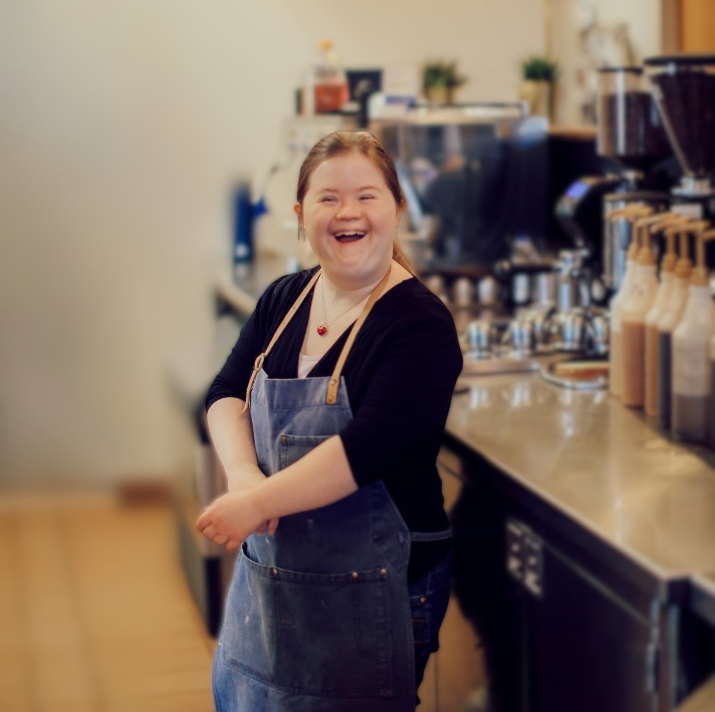 A woman with Down syndrome smiles at a coffee shop. She stands by the syrups and wears an apron.