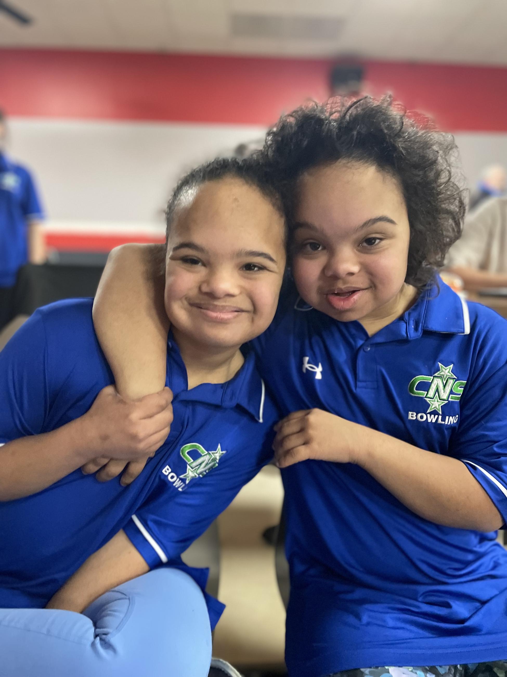 A girl with Down syndrome smiles with her sister. They both wear blue bowling shirts.