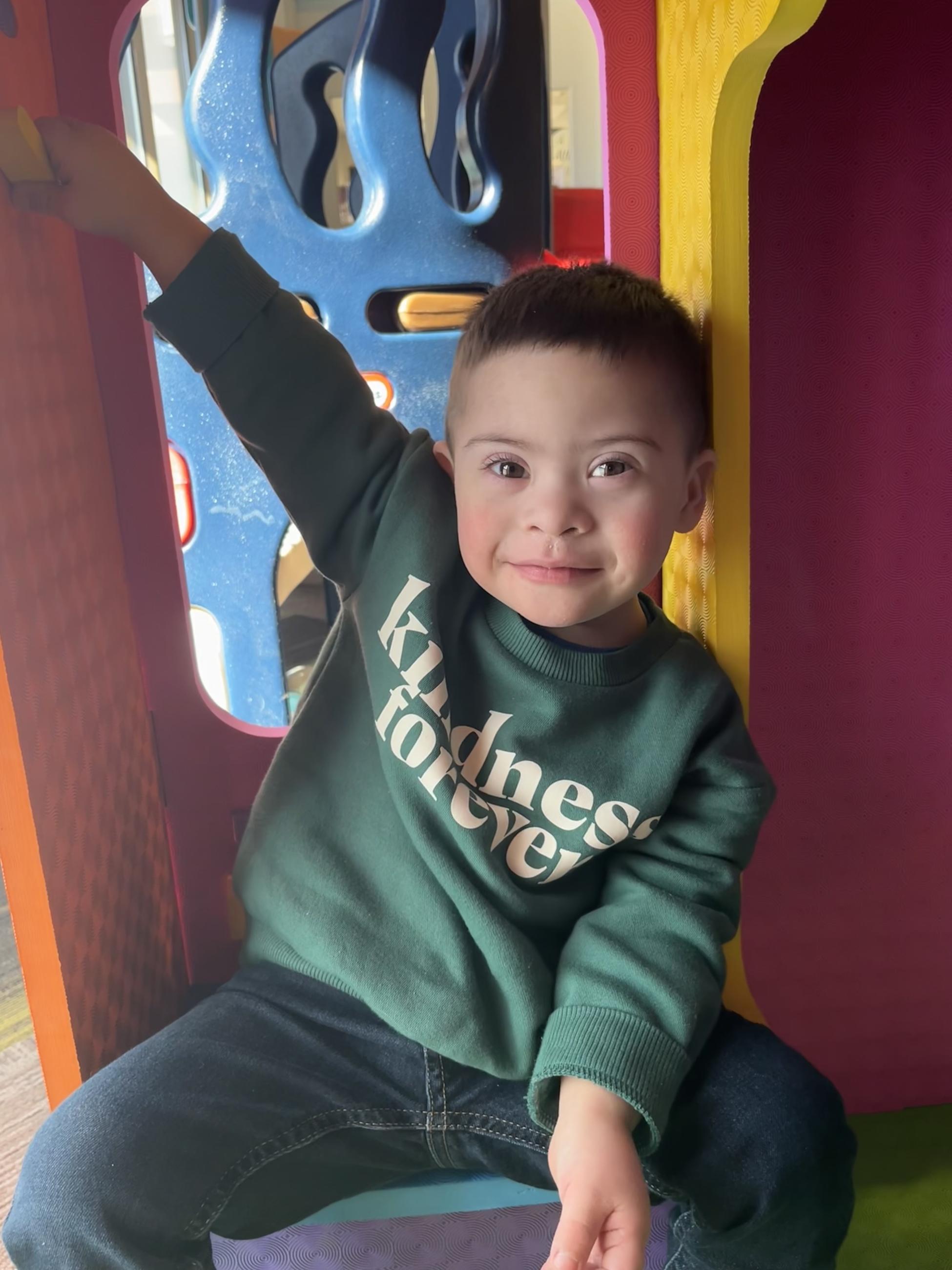 A young boy smiles at a playground. He wears a green shirt that says, "kindness forever"