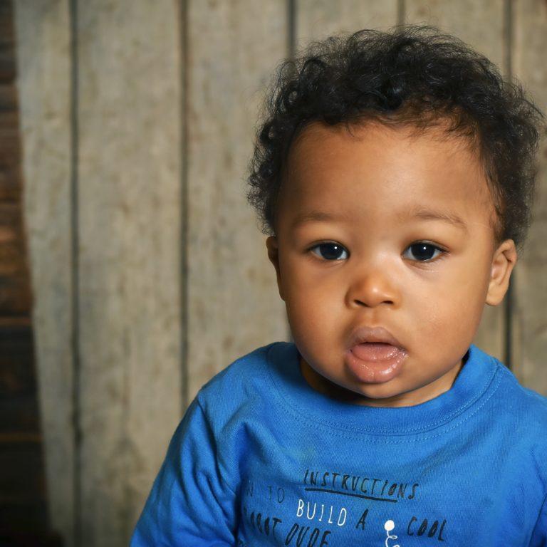 Image of a young toddler with Down syndrome. The toddler has curly black hair and wears a blue t-shirt.