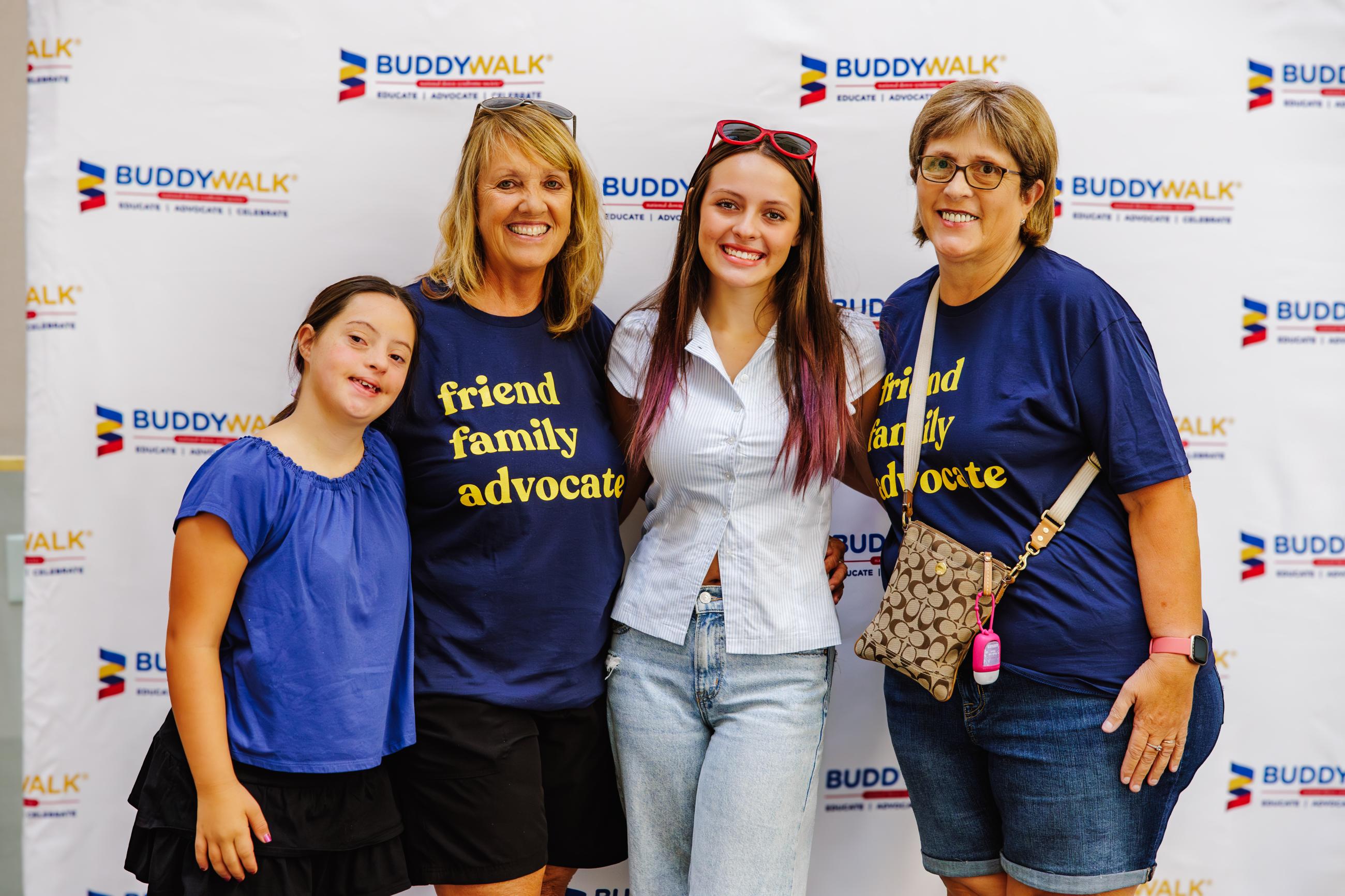 A family, including a girl with Down syndrome, pose in front of a Buddy Walk backdrop. Two of the women wear navy shirts with yellow text that say, "friend, family, advocate."