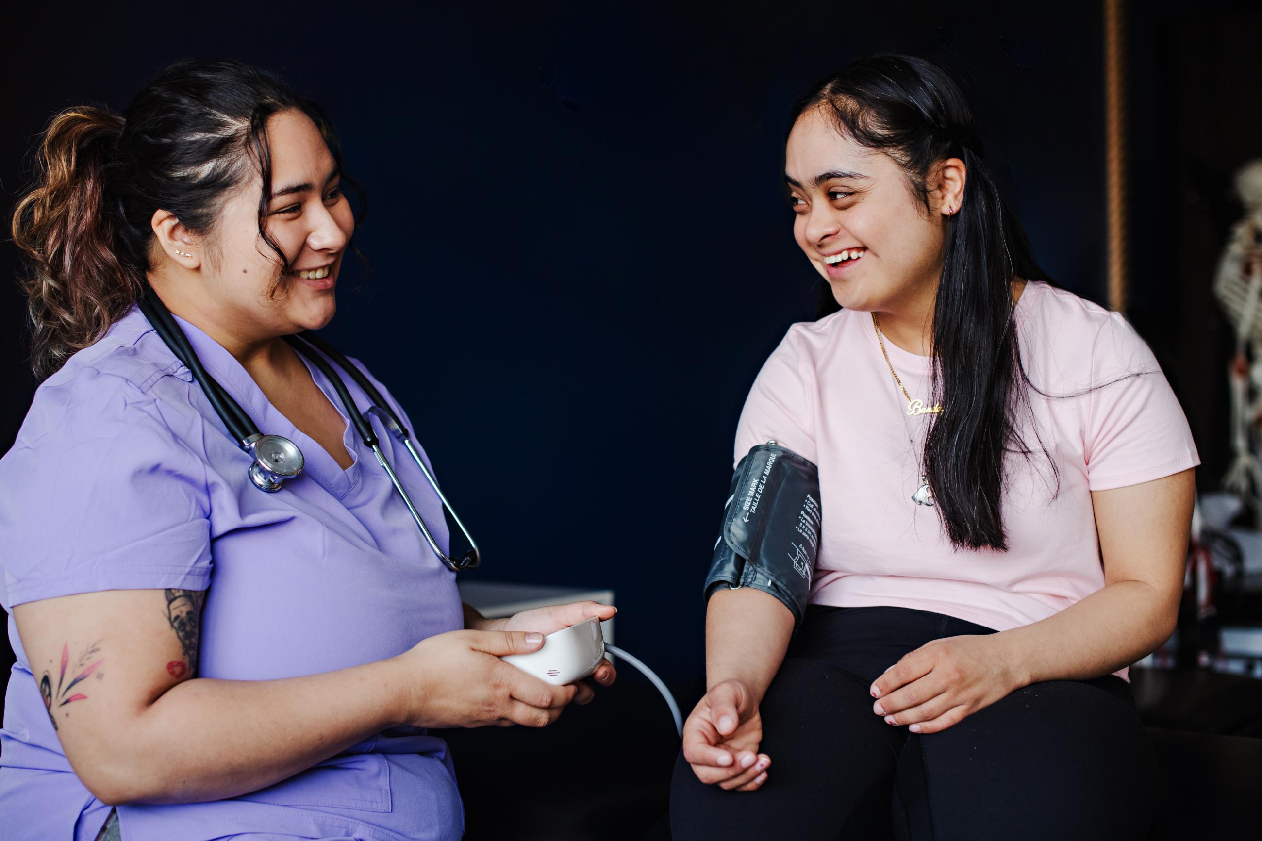 A female doctor in purple scrubs takes the blood pressure of a young girl in a pink shirt.