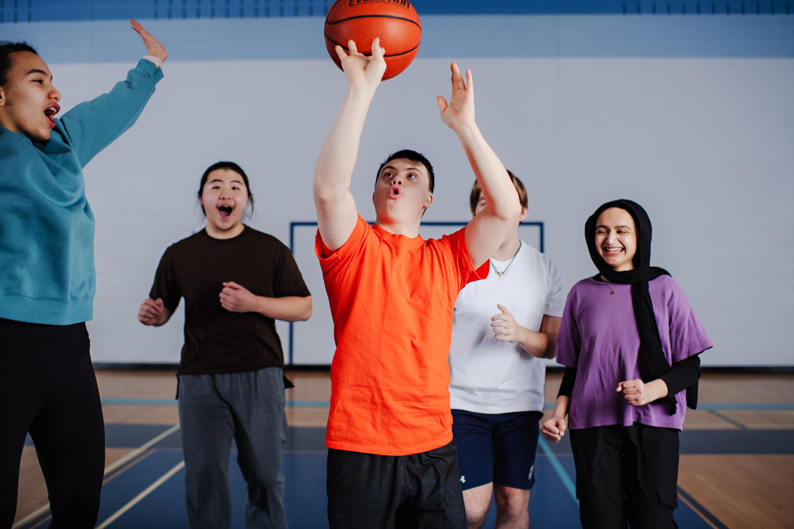A young man with Down syndrome shoots a basketball while playing indoors with a cheering group of friends.