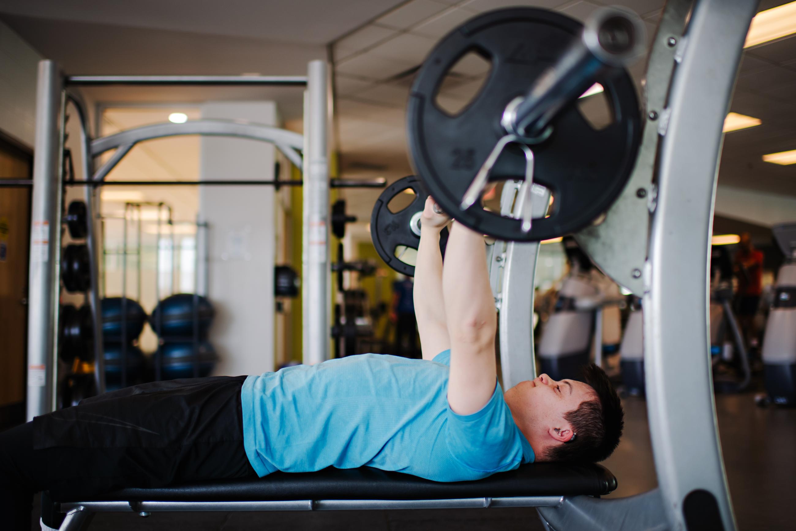 A young man with Down syndrome lifts a barbell while bench pressing at the gym.