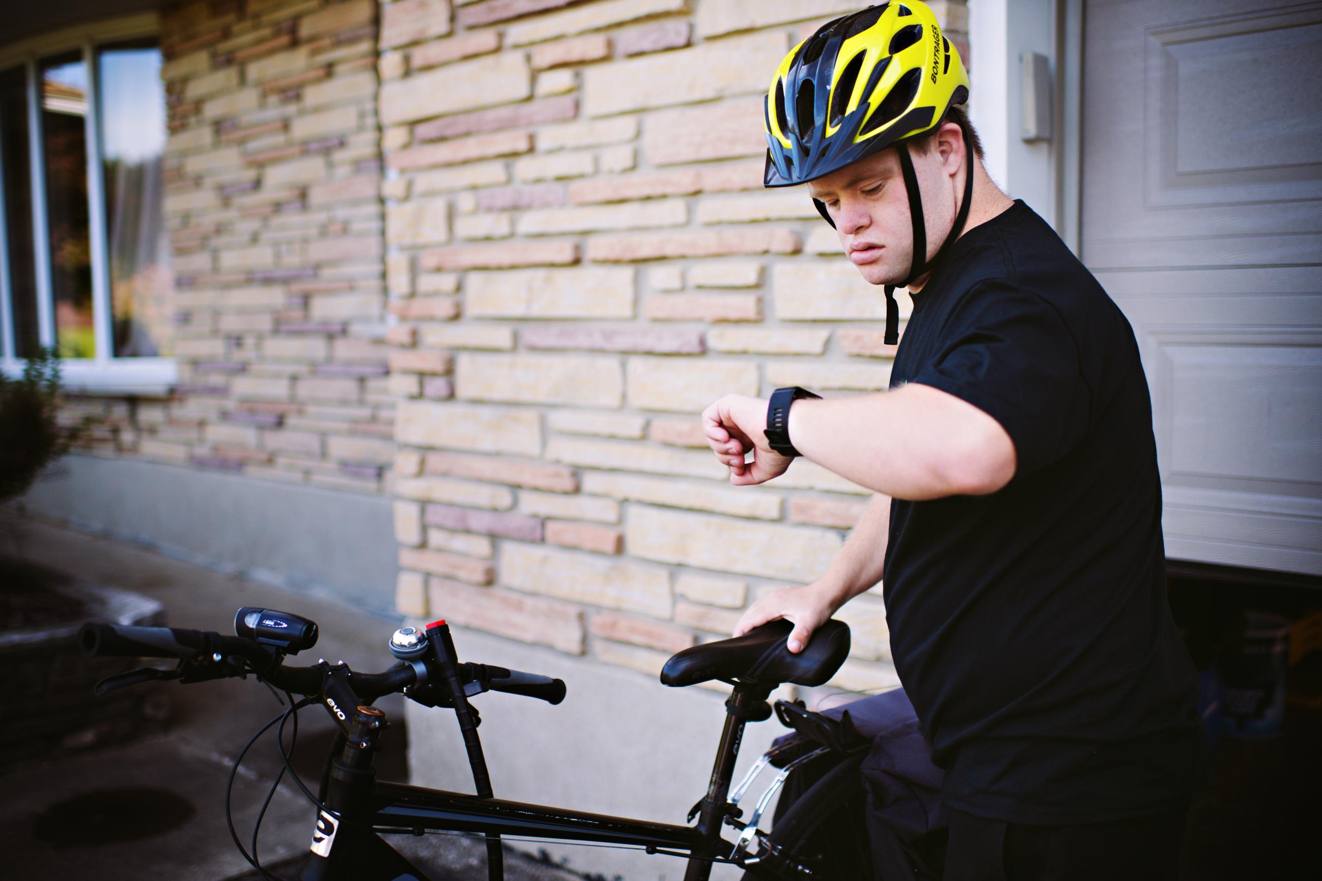 A young man with Down syndrome wearing a yellow helmet checks his smartwatch while standing next to his bicycle outside a home.