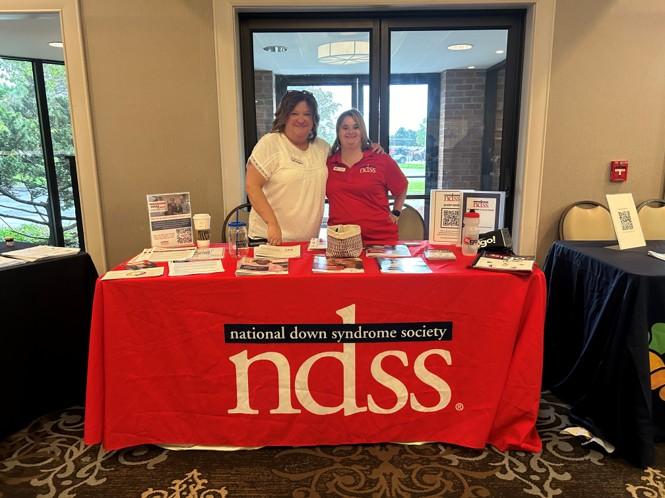 A woman with Down syndrome, wearing a red shirt, and a woman wearing a white shirt stand at a table with a red NDSS table cloth.