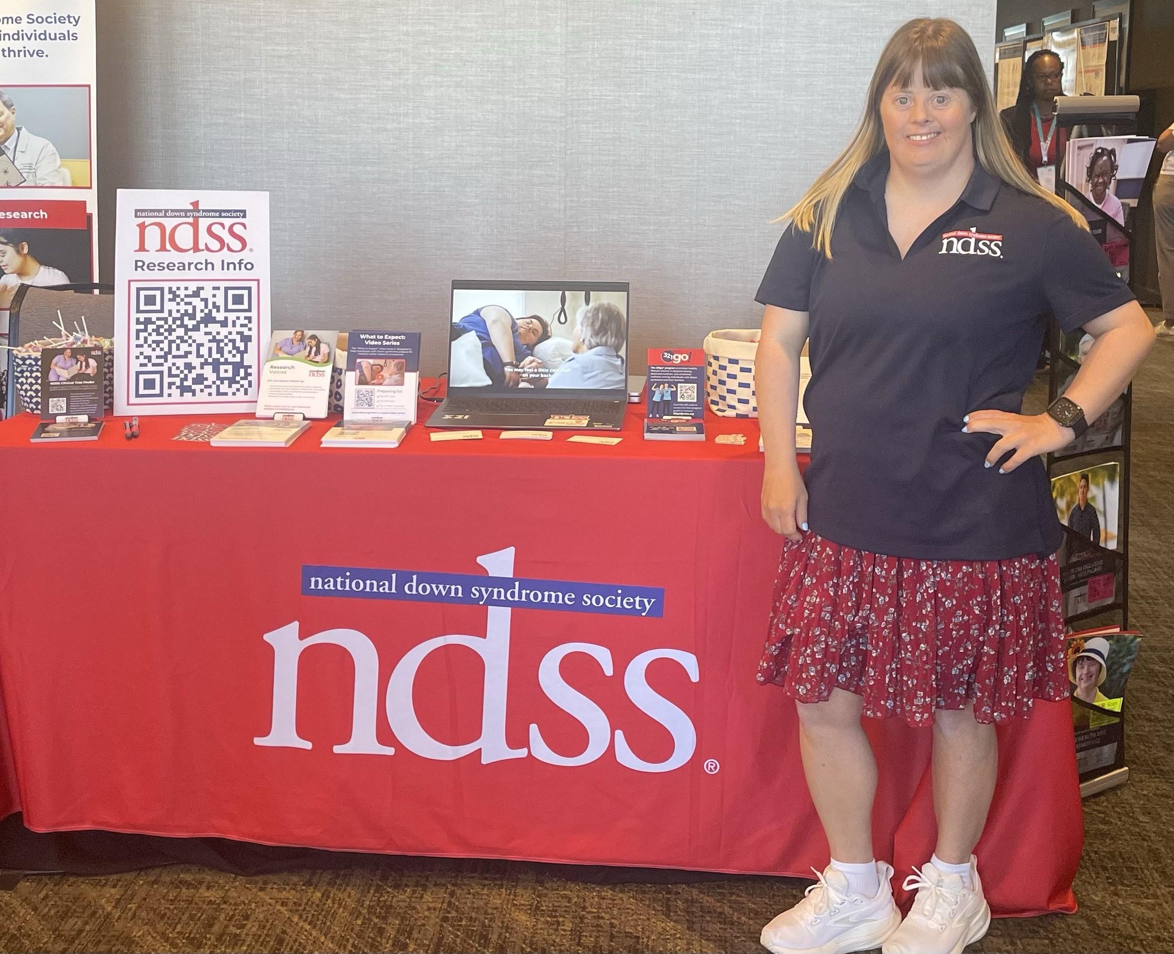 A woman with Down syndrome wears a polo shirt with the NDSS logo. She stands in front of an NDSS table with information about research.