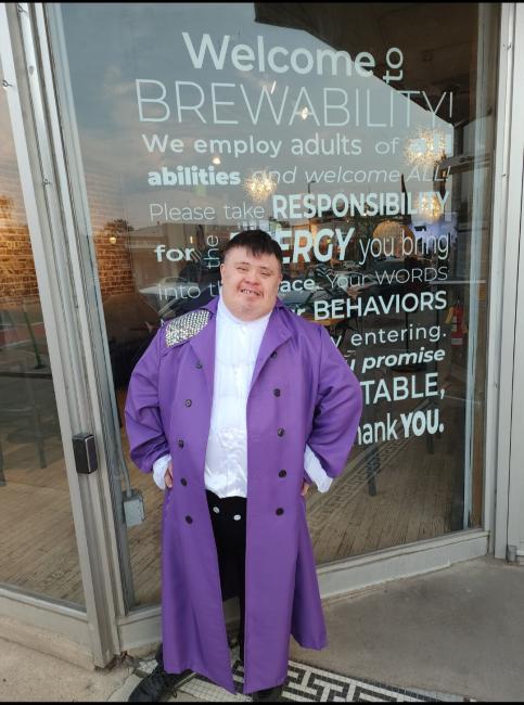 Man with Down syndrome smiles standing outside of his place of work, wearing a purple trench coat