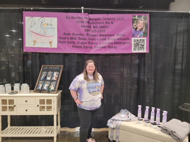 woman with down syndrome stands in front of her business at an expo, bath bombs