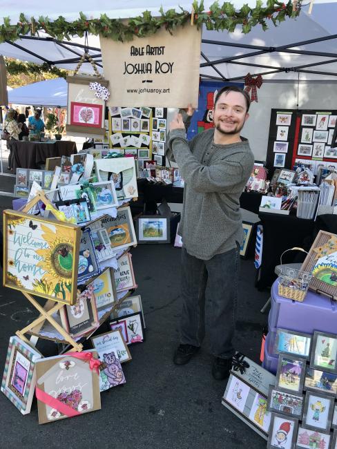 man with Down syndrome stands in front of his artistry at an open air market. he wears a grey sweater and black slacks