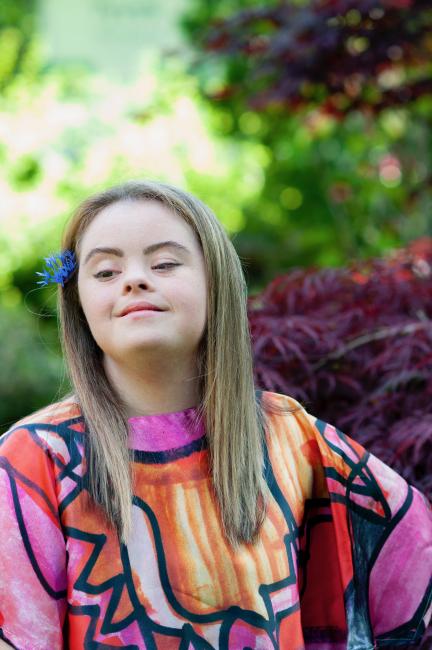 woman with down syndrome has brown hair and smiles in the distance wearing one of her original dress designs in red pink and orange designs