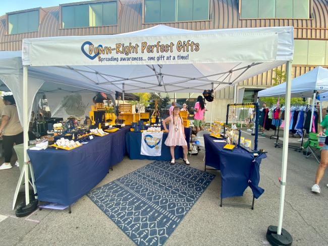 Hannah, a young white woman with Down syndrome, stands in front of her business booth. She wears a pink dress and her hair in two braids.