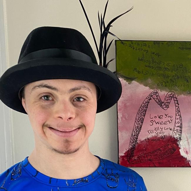 Joel, a man with Down syndrome who has a black hat, stands next to his artwork.