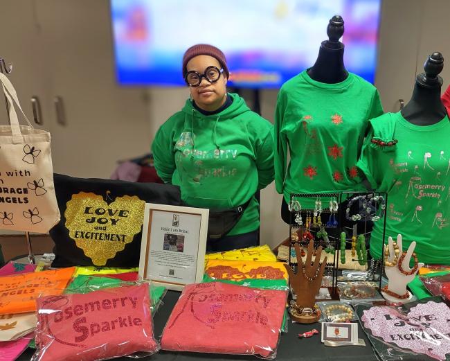 Britni, a woman with Down syndrome, wears round black glasses and a green sweatshirt. She stands behind her table of merchandise.