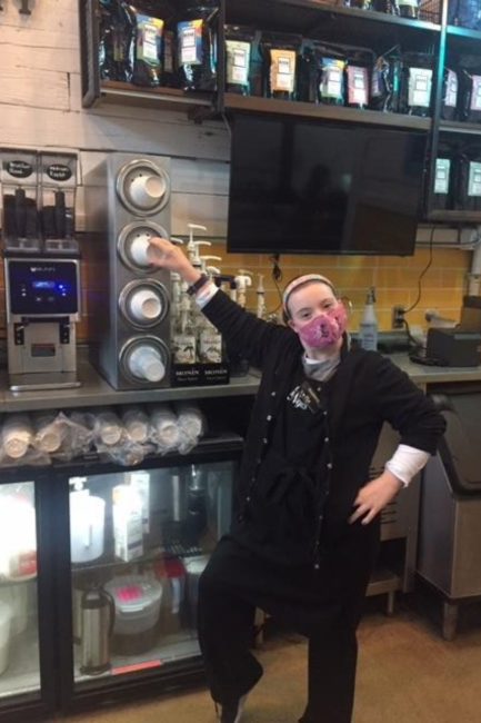 Kelley Rouse is wearing a face mask and pointing to an espresso maker at work