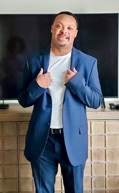 Sean Davis, a Black man with Down syndrome is smiling at the camera. He is wearing a white shirt and navy suit.