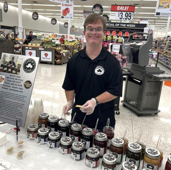 A man with Down syndrome, wearing glasses and a black Jared's Jams polo shirt, stands behind a sampling table in a grocery store. The table displays multiple jars of Jared's Jams products in various varieties, along with small tasting samples. A promotional sign displaying the Jared's Jams logo and business information for their Edina, Missouri location is visible beside the table. The grocery store's produce section and checkout area can be seen in the background.