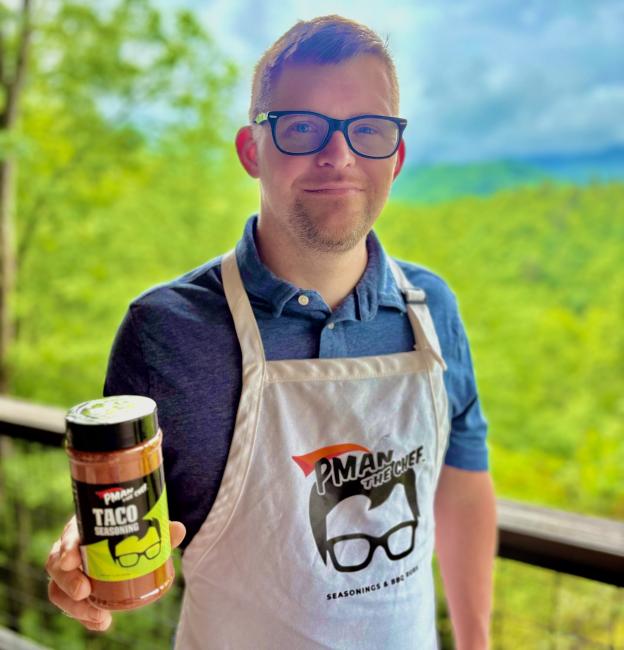 A man with Down syndrome, wearing glasses and a blue shirt stands outdoors against a scenic backdrop of green fields and blue sky. He's wearing a beige apron branded that has a Pman the Chef logo of an outline of a head with glasses, and holding a container of PMAN brand taco seasoning.