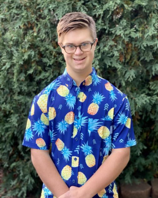 A young man with Down syndrome and light brown hair wears glasses and a Hawaiian t-shirt with pineapples on it. He poses in front of a large green plant outside.