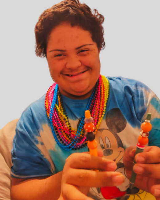 A young man with Down syndrome with short, curly auburn hair smiles at the camera against a light gray background. He wears a blue tie-dye T-shirt with a Mickey Mouse face and several layered necklaces of pink, blue, yellow, and orange beads, and holds up two beaded pens with orange beads.