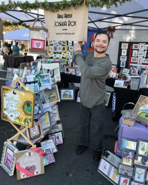 A man with Down syndrome and dark hair and a goatee smiles while holding up a tan fabric banner that reads “Able Artist Joshua Roy” and “www.joshuaroy.art” at an outdoor craft fair booth under a white canopy. He wears a gray long-sleeved sweater and dark jeans. Around him are framed artwork, greeting cards, and a wooden sign with sunflowers that reads “Welcome to our home.”