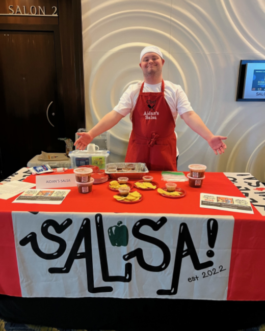 A man with Down syndrome, wearing a white cap and white T-shirt stands with arms outstretched and a big smile behind a table with a red tablecloth. His red apron reads “Chef Aidan’s Salsa” with a crossed-utensils icon. The table holds containers of salsa, bowls of tortilla chips, flyers, and a small sign reading “Aidan’s Salsa.” A banner across the front shows “Salsa!” in black lettering with a green bell pepper and “est 2022.” A sign on the wall behind reads “Salon 2.”