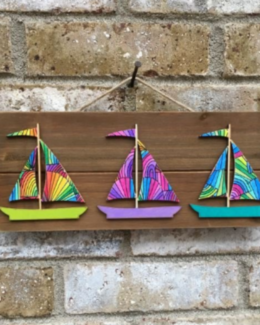 Wooden plank hangs by twine from a nail on a light brick wall. Three cutout wooden sailboats are attached to it, each with rainbow-patterned sails and a solid hull in green, purple, or teal.