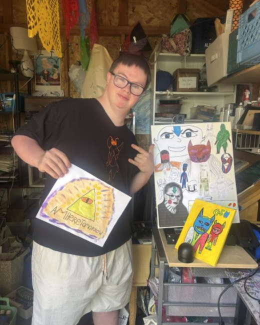 A man with Down syndrome with dark hair and dark-rimmed glasses tilts his head and holds up a drawing in a cluttered garage workspace. He wears a black T-shirt with an orange line drawing of a figure and light cream shorts. The drawing shows a yellow-green triangle with an orange eye inside, surrounded by yellow rays on an orange background, with the handwritten text “Am i here tomorrow” below. Behind him are a sketch of faces and creatures on white paper and a yellow canvas.