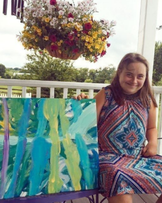 A woman with Down syndrome with long light brown hair smiles as she sits on a porch beside a large abstract painting in teal, green, yellow, and purple vertical brushstrokes. She wears a sleeveless dress with a blue, orange, and teal geometric pattern. A white porch railing, a hanging basket of red, yellow, and white flowers, and trees are visible behind her.