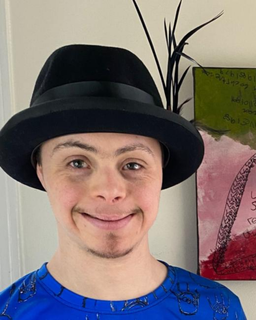 A smiling young man with Down syndrome and a light goatee looks straight at the camera. He wears a black felt fedora with a black band and a bright blue shirt with black hand-drawn line patterns. Behind him are a cream wall, a plant with long black feathers, and a painting in pink, red, and green with black line work.