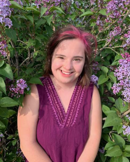 A smiling young woman with Down syndrome and shoulder-length brown hair with magenta streaks stands in front of a lilac bush with purple blossoms. She wears a sleeveless magenta V-neck top with embroidered trim.