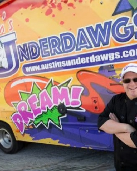 A man with Down syndrome, wearing sunglasses, a white cap, and a black chef’s jacket stands with his arms crossed and smiles beside a food truck painted in orange, yellow, and purple. The cropped truck lettering reads “Underdawgs” and “www.austinsunderdawgs.com,” with the word “Dream!” in pink letters on a green starburst.