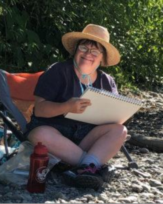 A smiling woman with Down syndrome in a straw hat and wire-rimmed glasses sits cross-legged in a camp chair on a rocky shore, holding a spiral sketchbook on her lap. She wears a dark purple short-sleeved top, dark shorts, gray socks, and hiking shoes. A red water bottle sits beside her, and green shrubs fill the background.