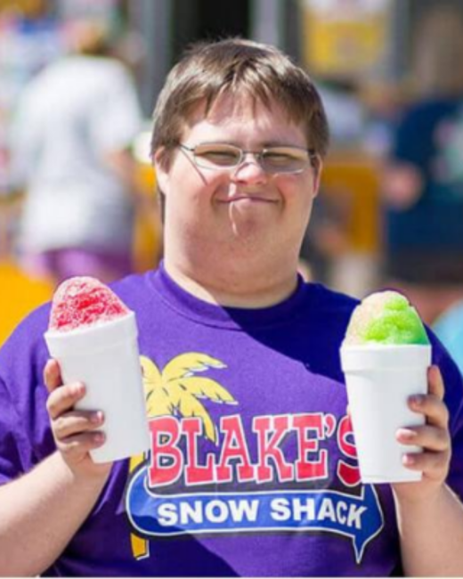 A man with Down syndrome, short brown hair, and wire-rimmed glasses smiles while holding two white foam cups of shaved ice, one red and one green. He wears a purple T-shirt with a yellow palm tree and the logo “Blake’s Snow Shack,” with “Blake’s” in red letters and “Snow Shack” in white letters on a blue banner. A blurred outdoor crowd is behind him.