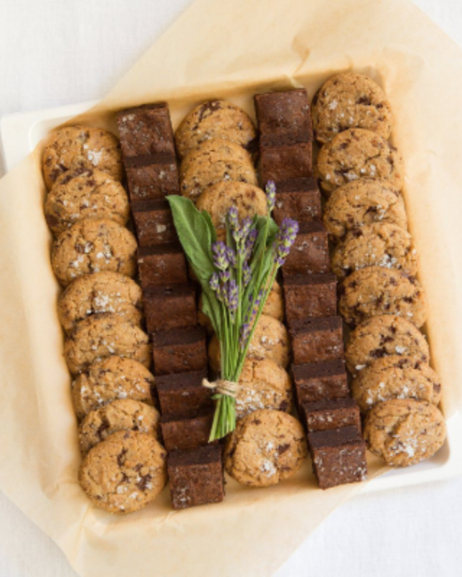 Overhead view of a white tray lined with parchment paper, holding alternating rows of salted chocolate chip cookies and brownie squares. A small bundle of lavender tied with twine lies in the center.
