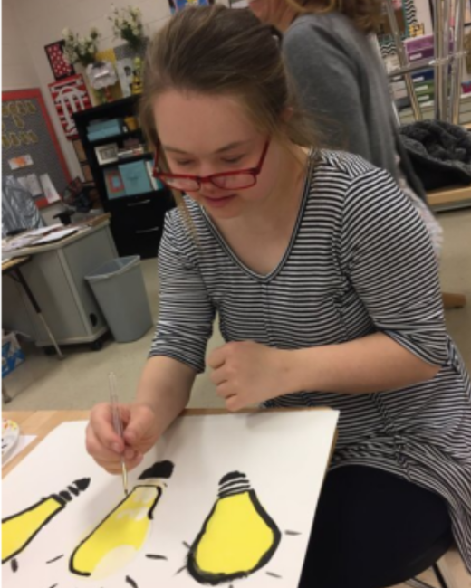 A young woman with Down syndrome, who has light brown hair pulled back and red-framed glasses, smiles slightly as she paints a picture of bright yellow light bulbs with a thin brush at a table in a classroom. She wears a black-and-white striped top. On the white paper in front of her are three yellow lightbulbs with thick black outlines, and a person in a gray cardigan stands behind her.