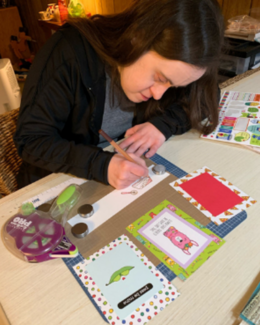A woman with Down syndrome and long dark brown hair looks down as she draws with a pencil at a wooden table. She wears a black jacket over a gray shirt. In front of her are handmade greeting cards, including one with a pink creature, one with a green pea pod, and one with a red square, along with a purple tape runner and a sheet of stickers.