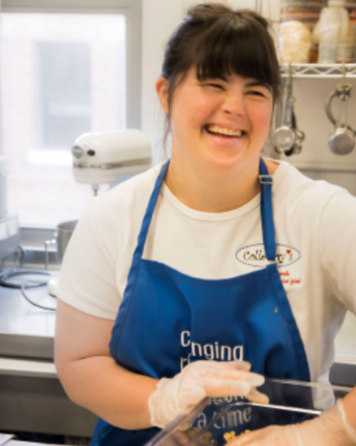 A woman with Down syndrome, dark brown hair, and bangs laughs while working in a commercial kitchen with a white stand mixer behind her. She wears a white T-shirt with an oval “Collettey’s” logo with a small red heart, a royal blue apron, and clear food-service gloves, and holds a clear container.