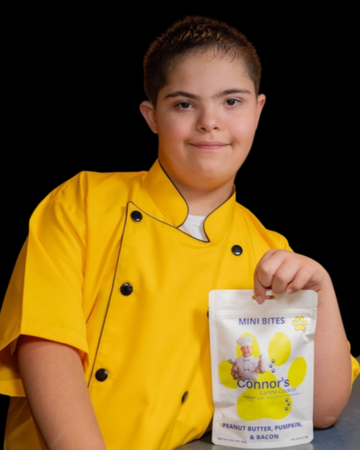 A young man with Down syndrome and short dark hair stands against a black background holding a white pouch of dog treats. He wears a bright yellow chef’s coat with black buttons and black trim. The pouch shows a yellow paw print, a small illustration of a chef, and the text “Mini Bites,” “Connor’s Canine Cookies,” and “Peanut butter, pumpkin, & bacon.”
