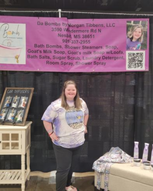 A woman with Down syndrome and long light brown hair stands smiling with one hand on her hip in a vendor booth with a black curtain backdrop. She wears a purple tie-dye T-shirt with a leopard-print graphic and black leggings. Behind her, a pink banner reads “Da Bombs by Morgan Tibbens, LLC, 3590 Windermere Rd N, Nesbit, MS 38651, 901-337-2955, Bath Bombs, Shower Steamers, Soap, Goat’s Milk Soap, Goat’s milk Soap w/Loofa, Bath Salts, Sugar Scrub, Laundry Detergent, Room Spray, Shower Spray.”
