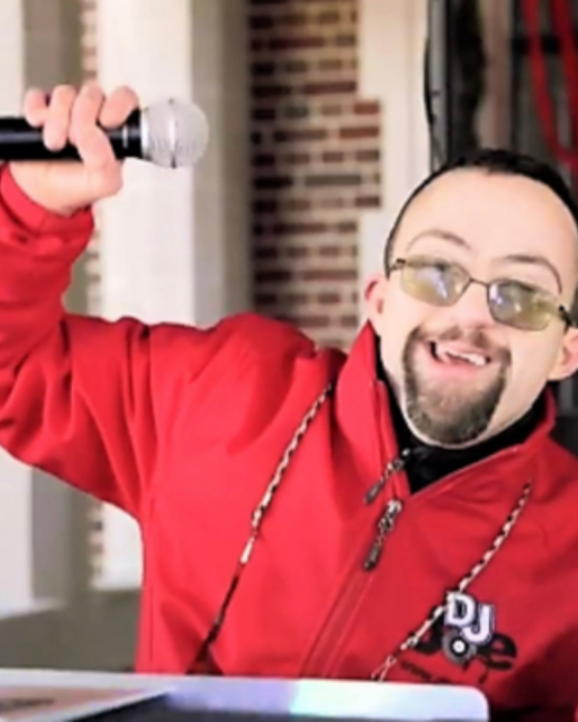 A man with Down syndrome, short dark hair, a goatee, and tinted glasses grins and raises a microphone in one hand. He wears a red zip-up jacket with a “DJ Joe” logo on the chest and red-and-white braided cords around his neck. A brick wall and white column are behind him.
