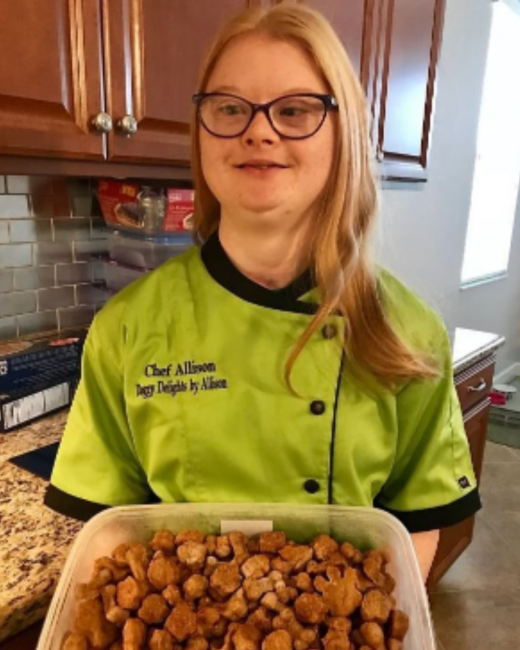 A woman with Down syndrome, long strawberry-blonde hair, and dark-rimmed glasses stands in a kitchen with brown cabinets and a granite countertop, holding a clear container of golden-brown dog treats. She wears a lime green chef’s coat with black trim and embroidered text that reads “Chef Allison, Doggy Delights by Allison.”