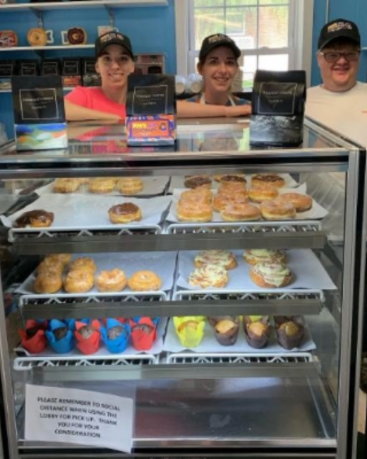 Three smiling people in black baseball caps stand behind a glass bakery display case: a young woman in a coral pink shirt, a young woman in a dark top and white apron, and a young man with Down syndrome in glasses and a white shirt. The case holds trays of glazed doughnuts, cinnamon rolls, and muffins in red, blue, and yellow liners, with bags of coffee on top and a blue wall behind.