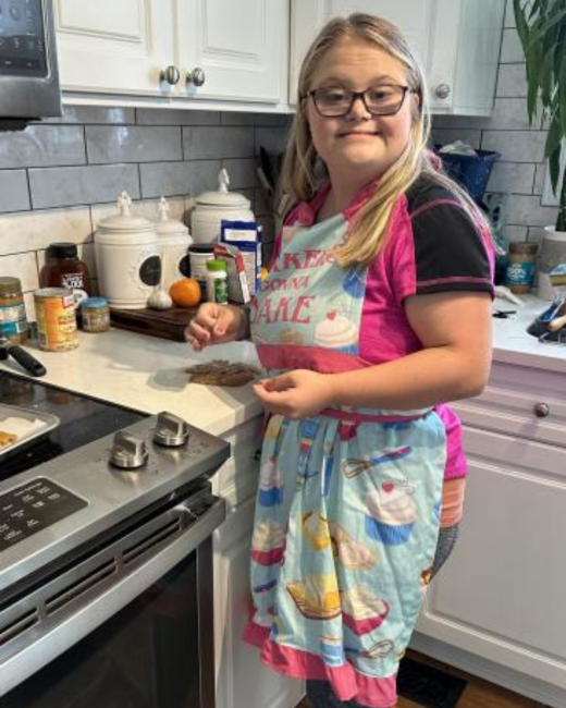 A young woman with Down syndrome, long blond hair, and dark-rimmed glasses smiles at the camera while standing at a kitchen counter with cookies on it. She wears a pink and black athletic shirt under a light blue apron decorated with cupcakes and whisks and printed with the words “Bakers gonna bake.” A stainless steel stove, white cabinets, and a gray tile backsplash are behind her.