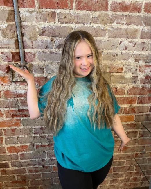 Smiling young woman with Down syndrome and long, wavy blond-highlighted hair poses with one hand resting on a red brick wall. She wears an oversized teal T-shirt and black leggings.