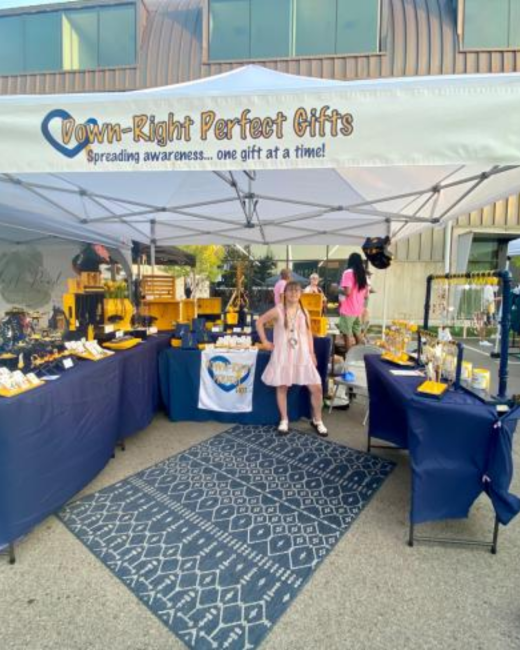 Outdoor vendor booth under a white canopy with a banner that reads “Down-Right Perfect Gifts” in gold letters beside a blue heart outline, with the tagline “Spreading awareness... one gift at a time!” Tables with navy cloths hold jewelry and gifts on yellow and black displays. A young woman with Down syndrome in a pink dress stands behind the center table beside a small white sign with the same logo, and a navy patterned rug covers the pavement in front.