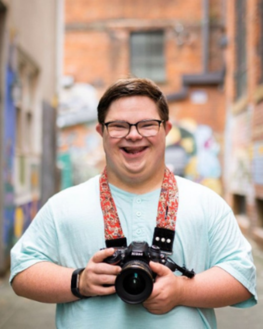 A young man with Down syndrome, short brown hair, and dark-rimmed glasses laughs as he holds a black DSLR camera in both hands. He wears a light mint T-shirt, a black wristband, and a red paisley camera strap, and stands in an alley with brick buildings and colorful murals behind him.