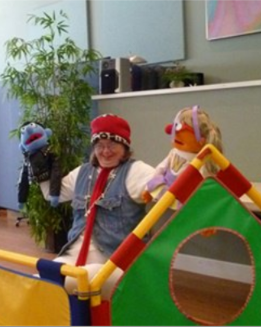 A woman with Down syndrome in glasses, a red knit hat, a denim vest over a white shirt, and a red scarf smiles while performing with two puppets: a blue furry puppet on one raised hand and a puppet with an orange face, blond hair, and a lavender headband on the other. A colorful play structure in red, yellow, green, and blue sits in the foreground, with a leafy plant and shelves behind.